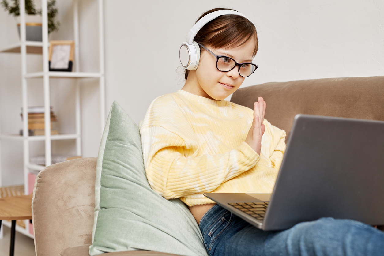 Young girl wearing eyeglasses and headphone making hand gestures in front of a laptop at home.